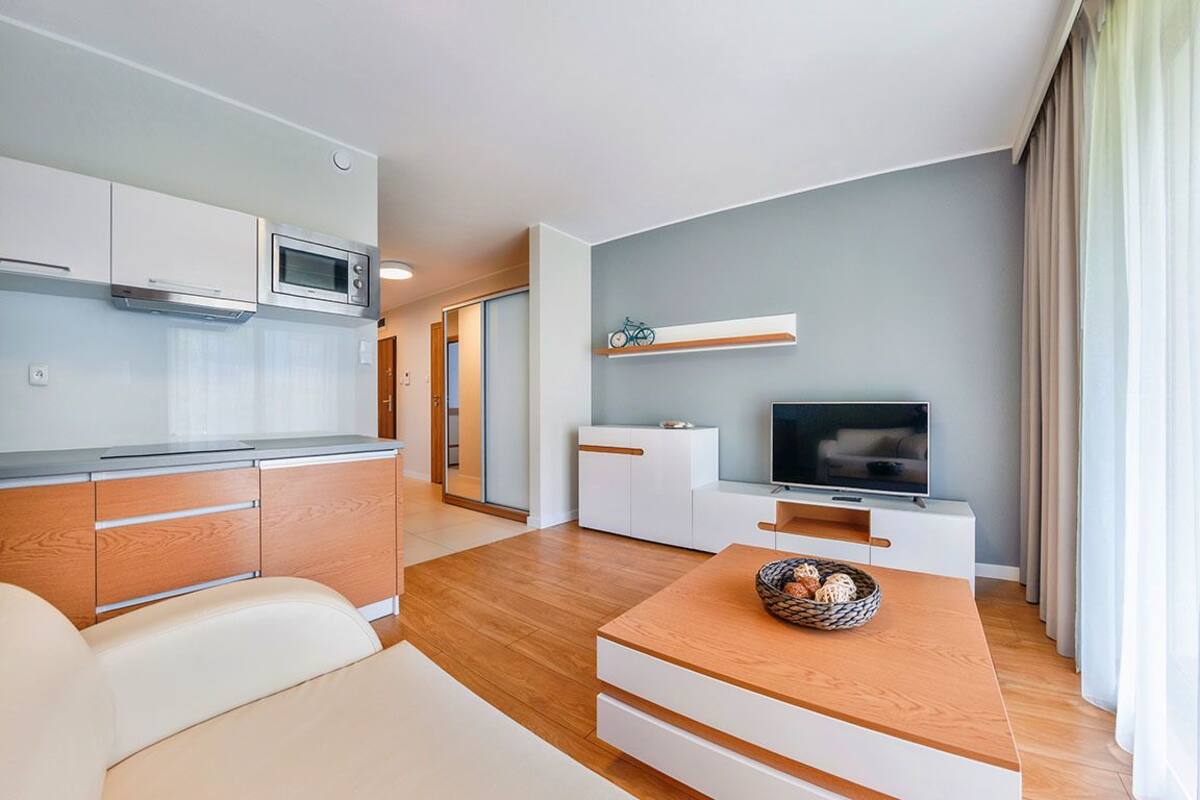 A modern living room with a wooden coffee table, a television, and a kitchen area with white cabinets and a microwave.
