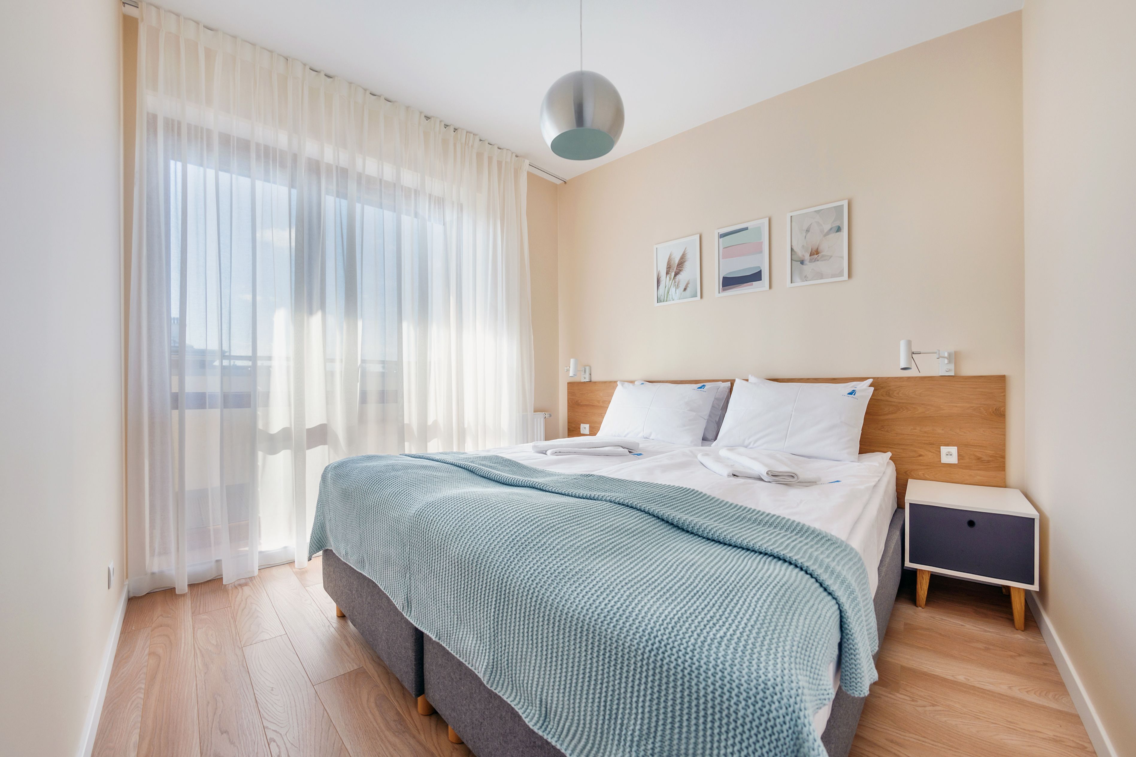 A bedroom with a bed, a bedside table, a nightstand, a window with sheer curtains, and wall art.