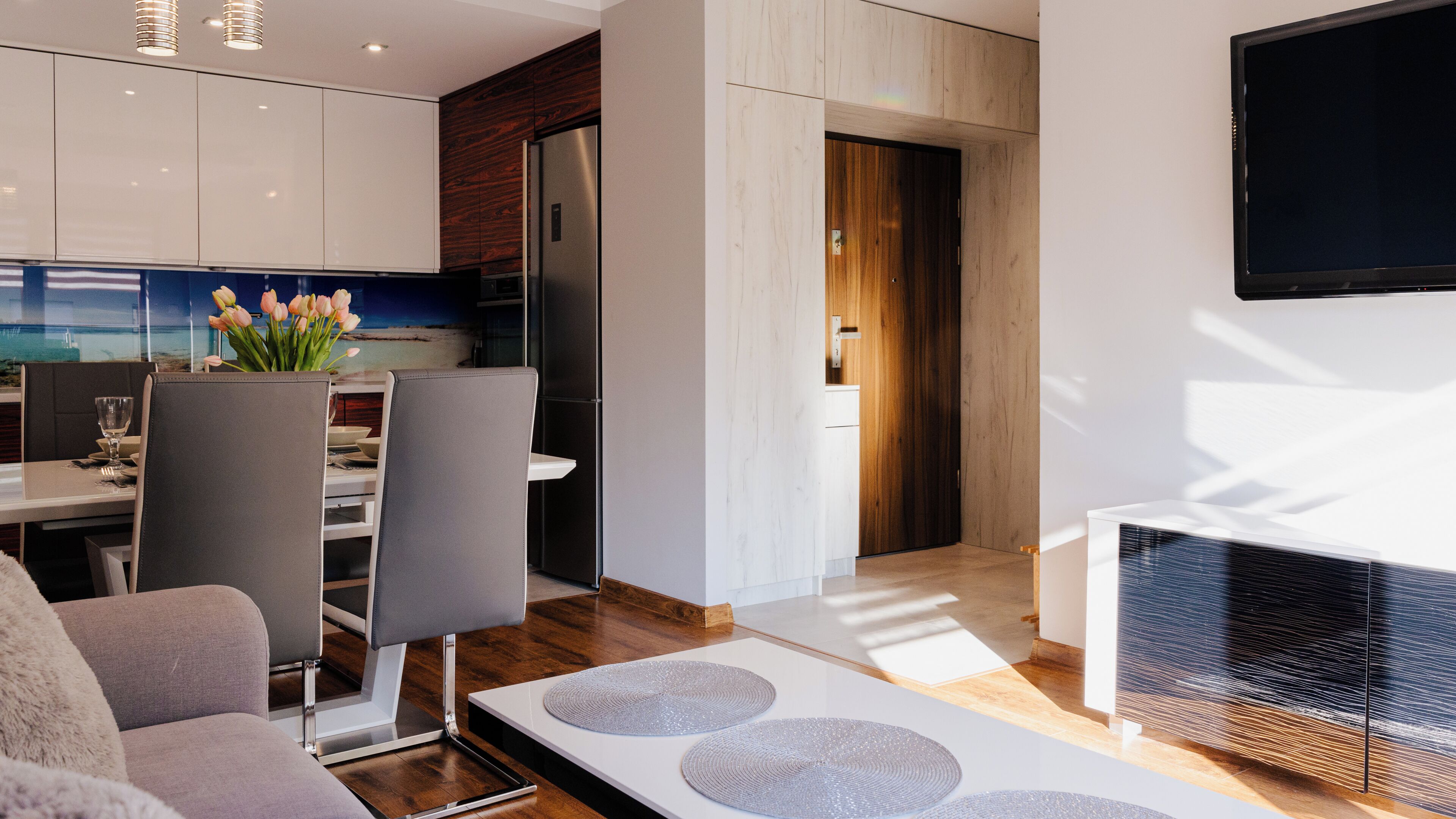A modern living room with a sofa, a dining table with chairs, and a kitchen area with a refrigerator and a TV mounted on the wall.