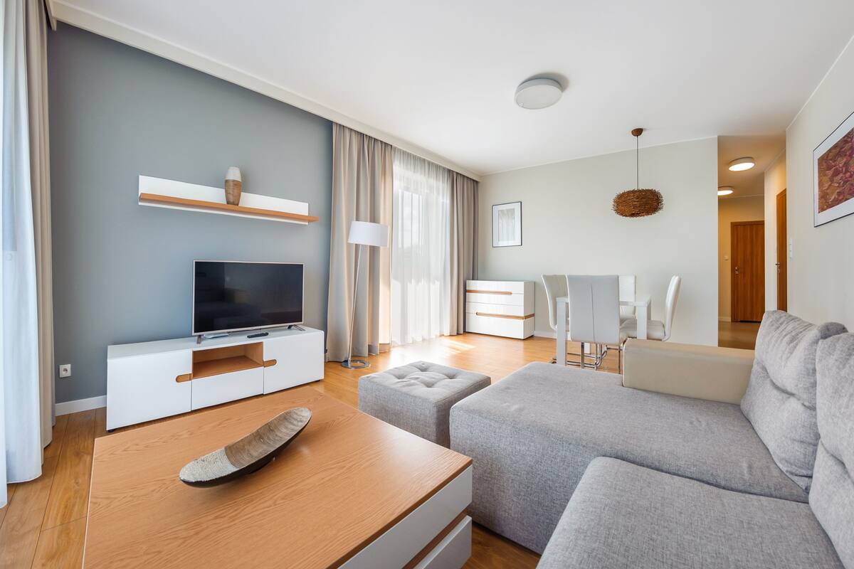 A modern living room with a flat-screen TV, grey sofa, wooden coffee table, and a dining area with a white table and chairs.