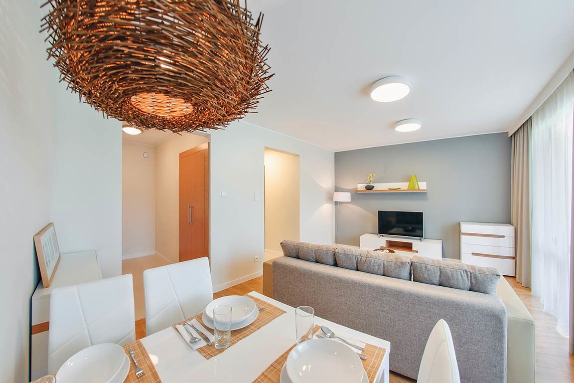 Una sala moderna con un sofá gris, un comedor con sillas blancas y una mesa para dos, y un televisor sobre un soporte blanco.
