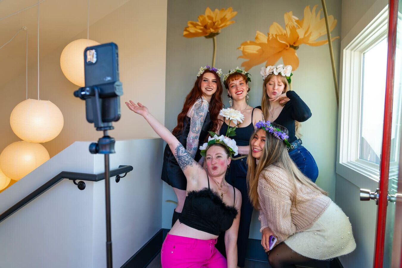 A group of people in a room with a staircase, a selfie stick, and decorative flowers.