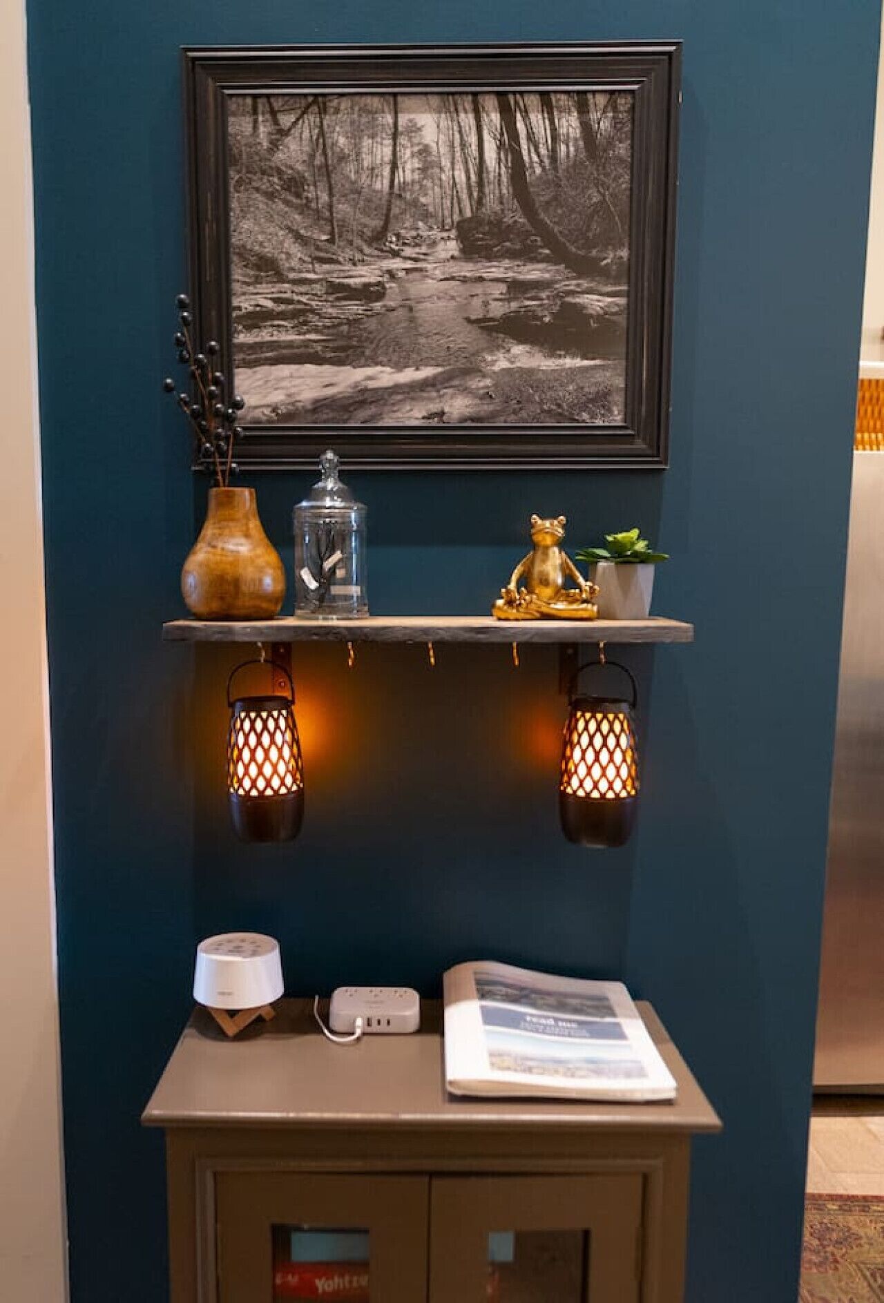 A shelf with a framed photograph, a vase, a glass jar, a frog figurine, and two lit lanterns. Below, there's a small table with a lamp, a power strip, and a book.