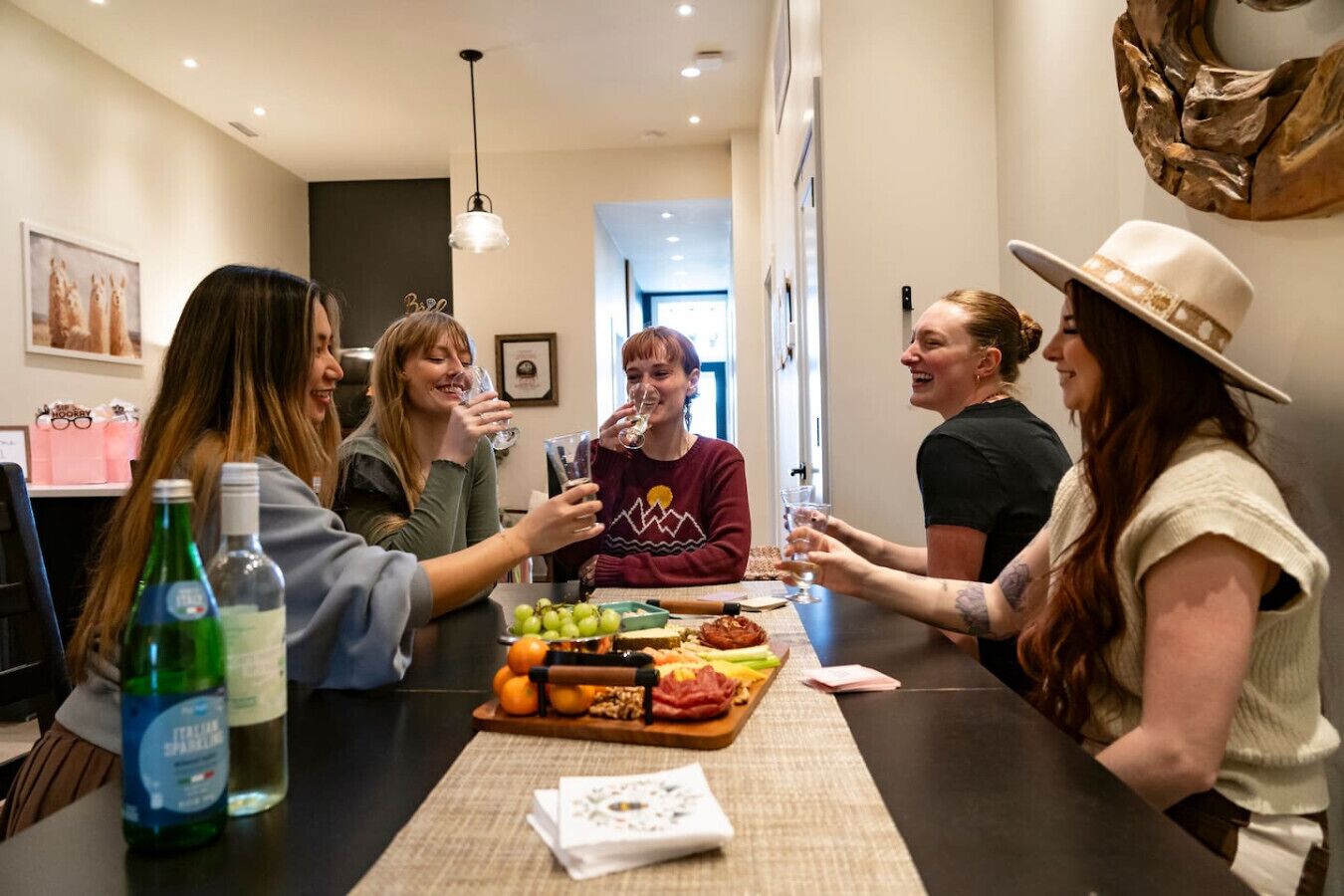 A group of people toasting at a table with a variety of food and drinks, in a modern interior with a kitchen visible in the background.
