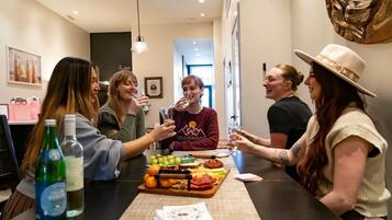 A group of people toasting at a table with a variety of food and drinks, in a modern interior with a kitchen visible in the background.