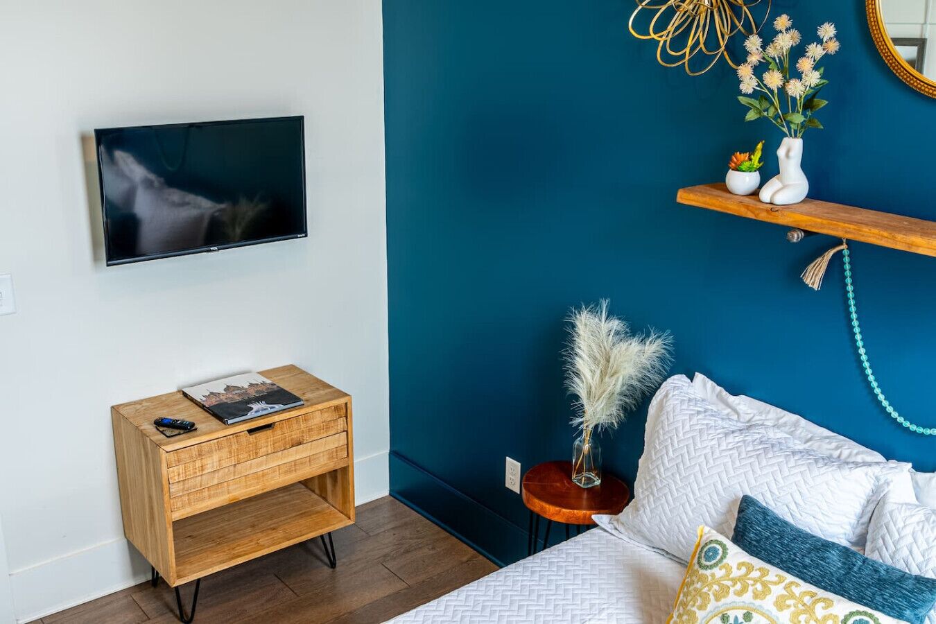 Une chambre à coucher comprenant un lit, un téléviseur fixé au mur, une table de chevet en bois, une petite table d’appoint et une étagère avec des objets décoratifs.