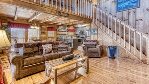 A cabin interior with a wooden staircase, a sofa set, a dining table, and a kitchen area.