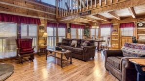 A spacious living room with a high ceiling, wooden beams, and a cozy fireplace. There is a comfortable seating area with a sofa and armchairs, and a dining table with chairs.