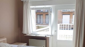 A bedroom with two beds, a window with curtains, a radiator, and a view of a building outside. - Appartment with seaside view and 2 parking spaces (Oostduinkerke-Bad)