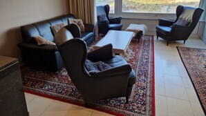 A living room with a black leather sofa, two armchairs, a coffee table, and a large window offering a view of the beach and dunes. - Appartment with seaside view and 2 parking spaces (Oostduinkerke-Bad)