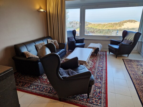 A living room with a black leather sofa, two armchairs, a coffee table, and a large window offering a view of the beach and dunes.