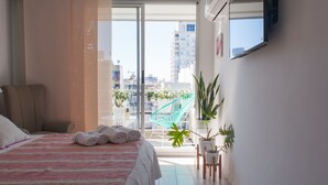 A modern hotel room with a large bed, a view of the city, and a balcony with plants.