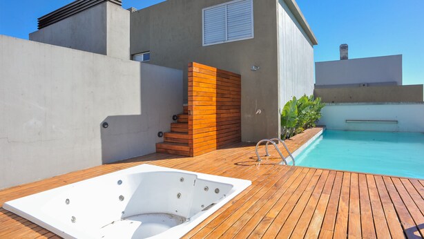 A modern outdoor pool area with a hot tub, wooden deck, and a concrete building with a window.
