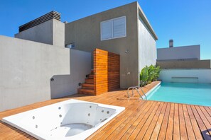 A modern outdoor pool area with a hot tub, wooden deck, and a concrete building with a window.