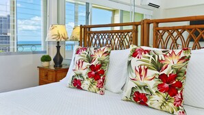 A hotel room with a bed, floral pillows, a nightstand with a lamp, and a view of the ocean through large windows. - Ocean & Sunset Views, FREE Parking & Washlet! (Honolulu)