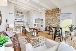 A spacious living area with a high ceiling, a stone wall, a wooden coffee table, leather sofas, and a TV mounted on the wall.