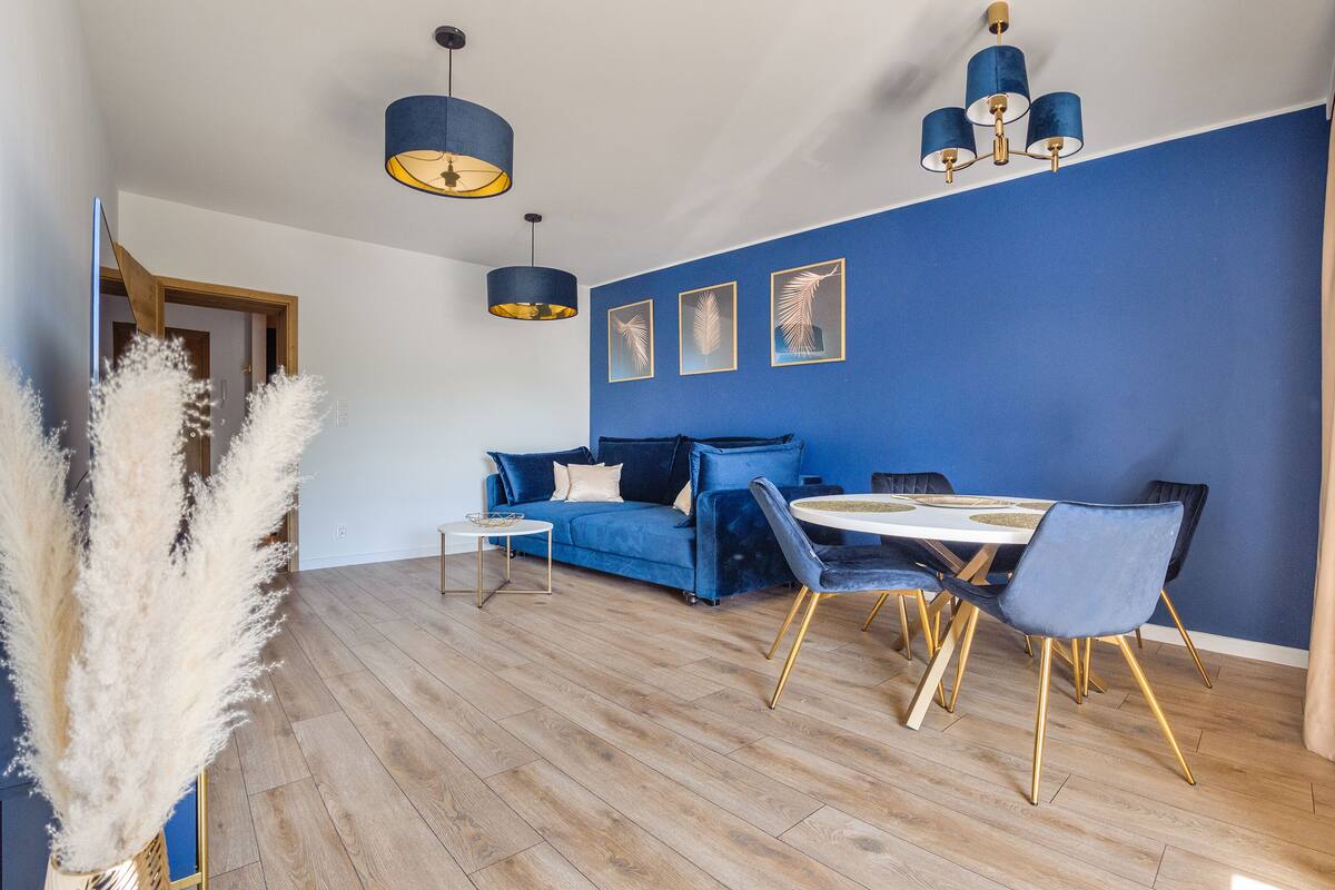 Un salon moderne avec un mur d’accent bleu, un plancher en bois, un canapé bleu, une table à manger avec des chaises et des luminaires suspendus.