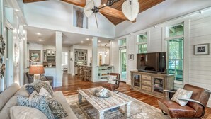 A spacious living room with a large sofa, a coffee table, and a leather armchair. There is a kitchen in the background with a dining area and a television.