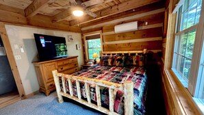 Habitació d'una cabana acollidora, amb sostre de fusta, un llit amb llençols de colors, un televisor sobre una petita taula de fusta, i una finestra amb vista a arbres.