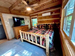 A cozy cabin room with a wooden ceiling, a bed with a colorful quilt, a TV on a wooden dresser, and a window with a view of trees. - 3BR_3BA Log Cabin, Pond Front, Hot Tub, Pool Table, near Skiing & Pet friendly (Seven Devils)