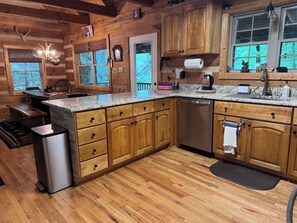 A wooden cabin kitchen with a central island, wooden cabinets, a stainless steel dishwasher, and a window overlooking greenery. - 3BR_3BA Log Cabin, Pond Front, Hot Tub, Pool Table, near Skiing & Pet friendly (Seven Devils)