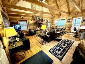 A cozy log cabin interior with a fireplace, wooden beams, a television, and a dining area. - 3BR_3BA Log Cabin, Pond Front, Hot Tub, Pool Table, near Skiing & Pet friendly (Seven Devils)