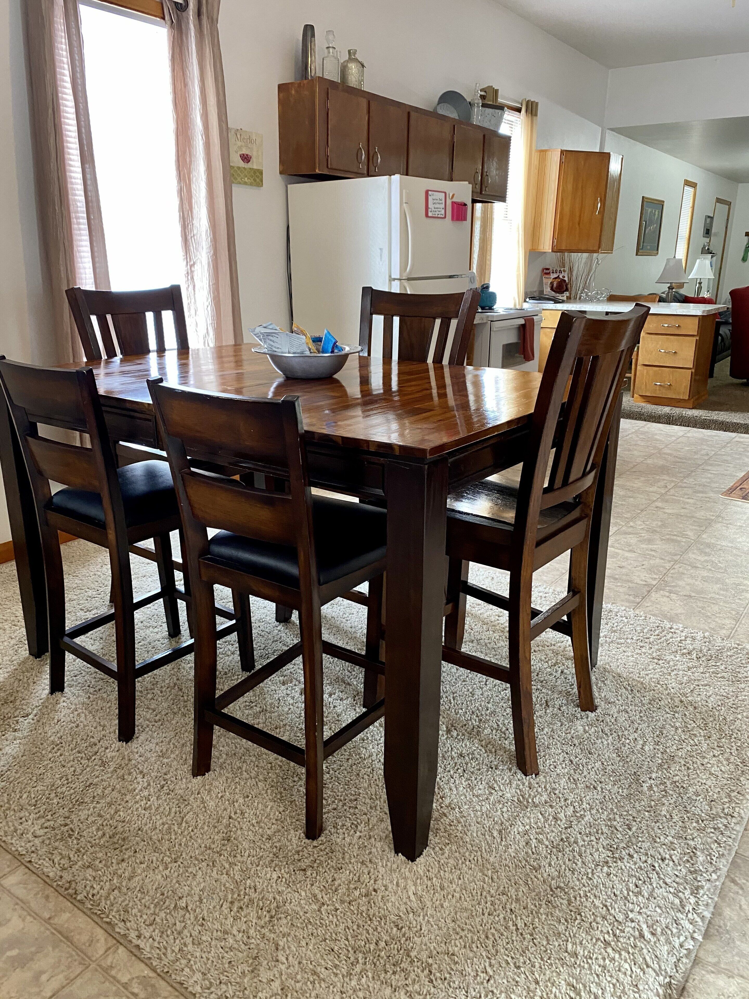A dining area with a wooden table and chairs, a kitchen in the background, and a rug on the floor.