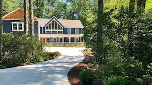 A two-story house with a covered porch, surrounded by dense greenery and trees. - Beautiful new large family home inside gates of Country Club of Sapphire Valley. (Sapphire)