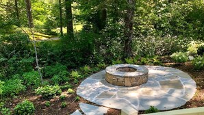A stone fire pit surrounded by a circular stone patio, with a wooden deck in the foreground. - Beautiful new large family home inside gates of Country Club of Sapphire Valley. (Sapphire)
