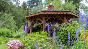 Un belvédère en bois, entouré d’un jardin luxuriant où s’épanouissent de nombreuses fleurs et plantes vertes.