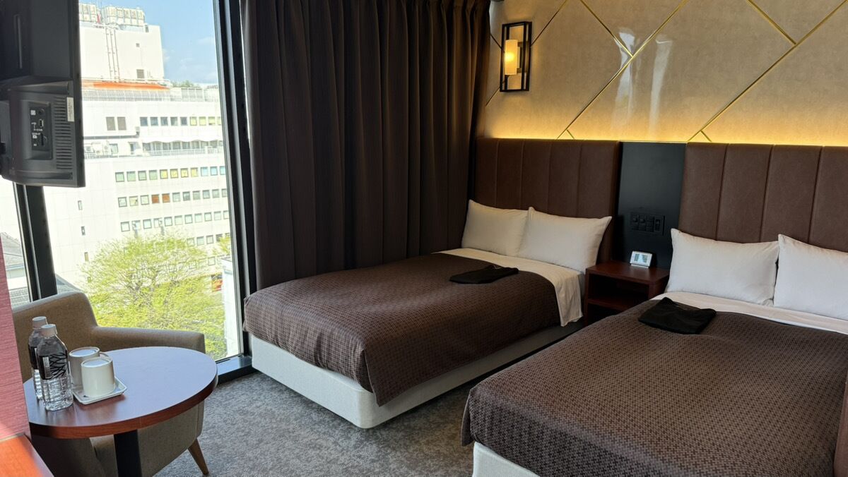 Habitación de hotel con dos camas, una mesita con una botella y una vela, un televisor y una ventana con vistas a edificios y árboles.