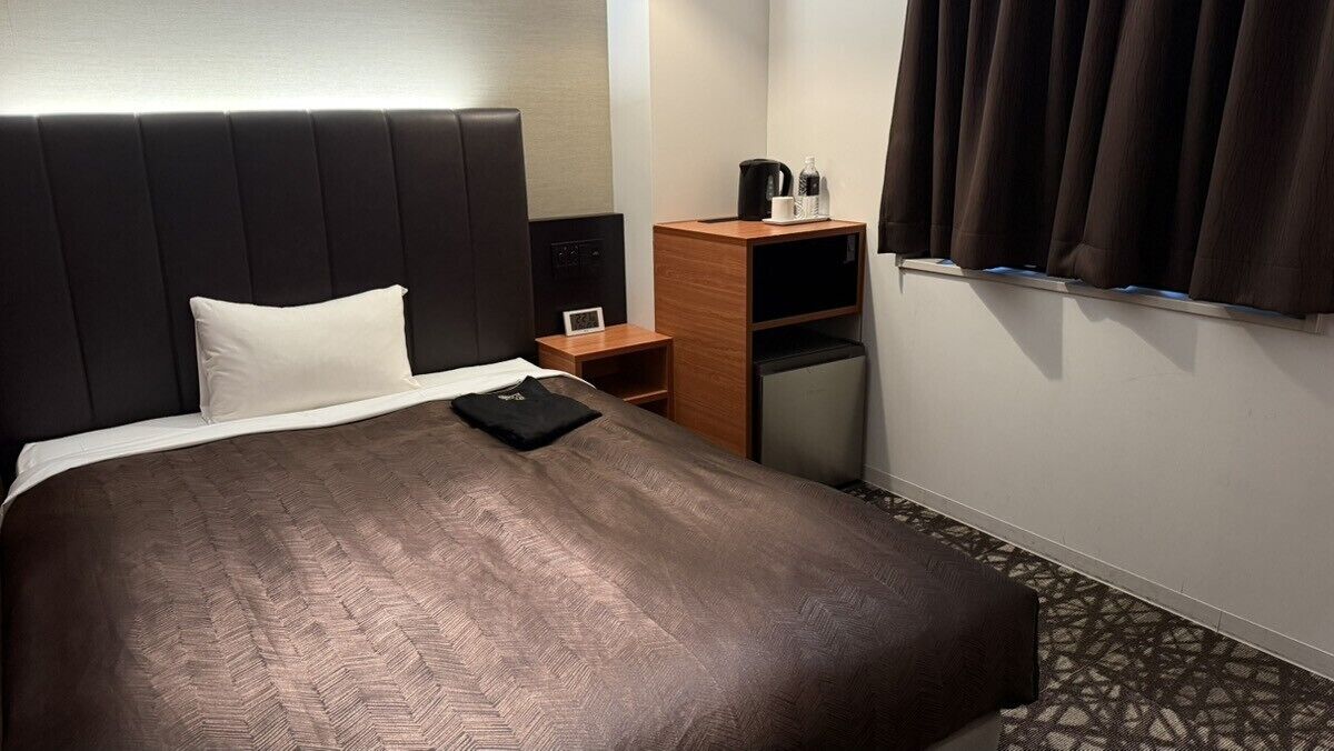 Habitación de hotel con una cama grande, una mesita de noche, un armario de madera, un refrigerador pequeño y una ventana con cortinas.