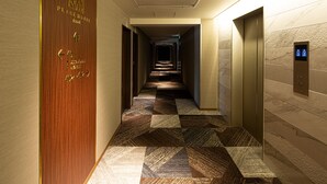 A hotel corridor with a wooden door on the left, a patterned carpet, and an elevator on the right. - PLACE Hotel Ascot (Kumamoto)