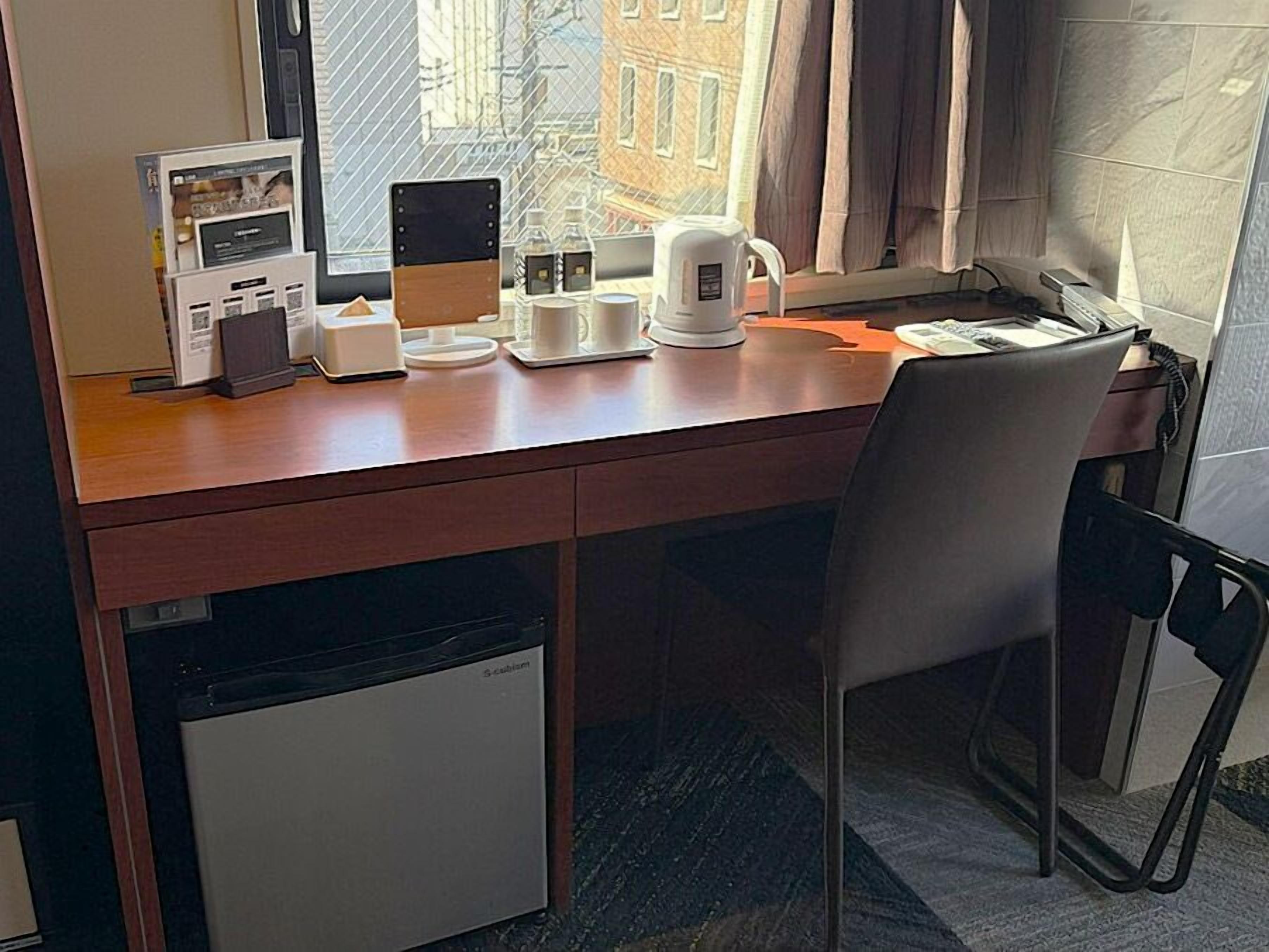 A hotel room with a wooden desk, a chair, a small refrigerator, a window with a view of buildings, and a coffee maker.