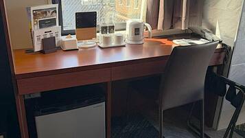 A hotel room with a wooden desk, a chair, a small refrigerator, a window with a view of buildings, and a coffee maker.