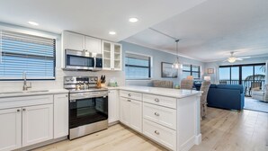 Een moderne keuken met witte kasten, een ingebouwde oven en een magnetron. Er is een zithoek met een blauwe bank en een rieten stoel.