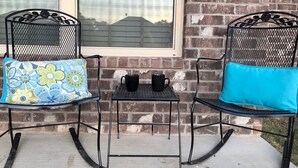 Een terras met twee zwarte metalen stoelen, waarvan er één een bloemenkussen en de ander een effen blauw kussen heeft, en een klein tafeltje met twee zwarte mokken.