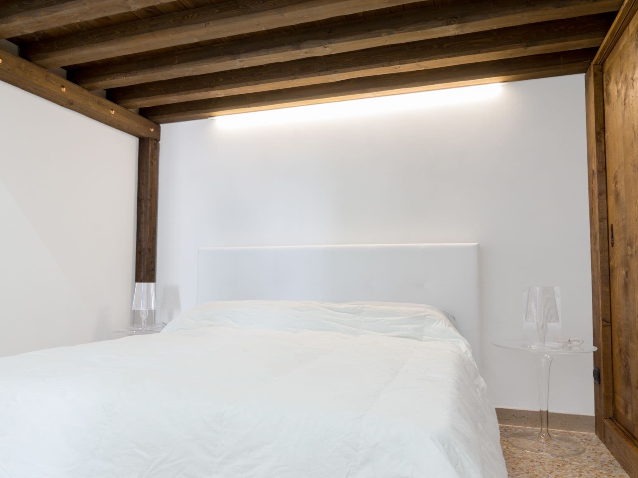Une chambre avec un plafond en bois, un lit doté d’une tête de lit blanche et une petite table avec un plateau en verre.