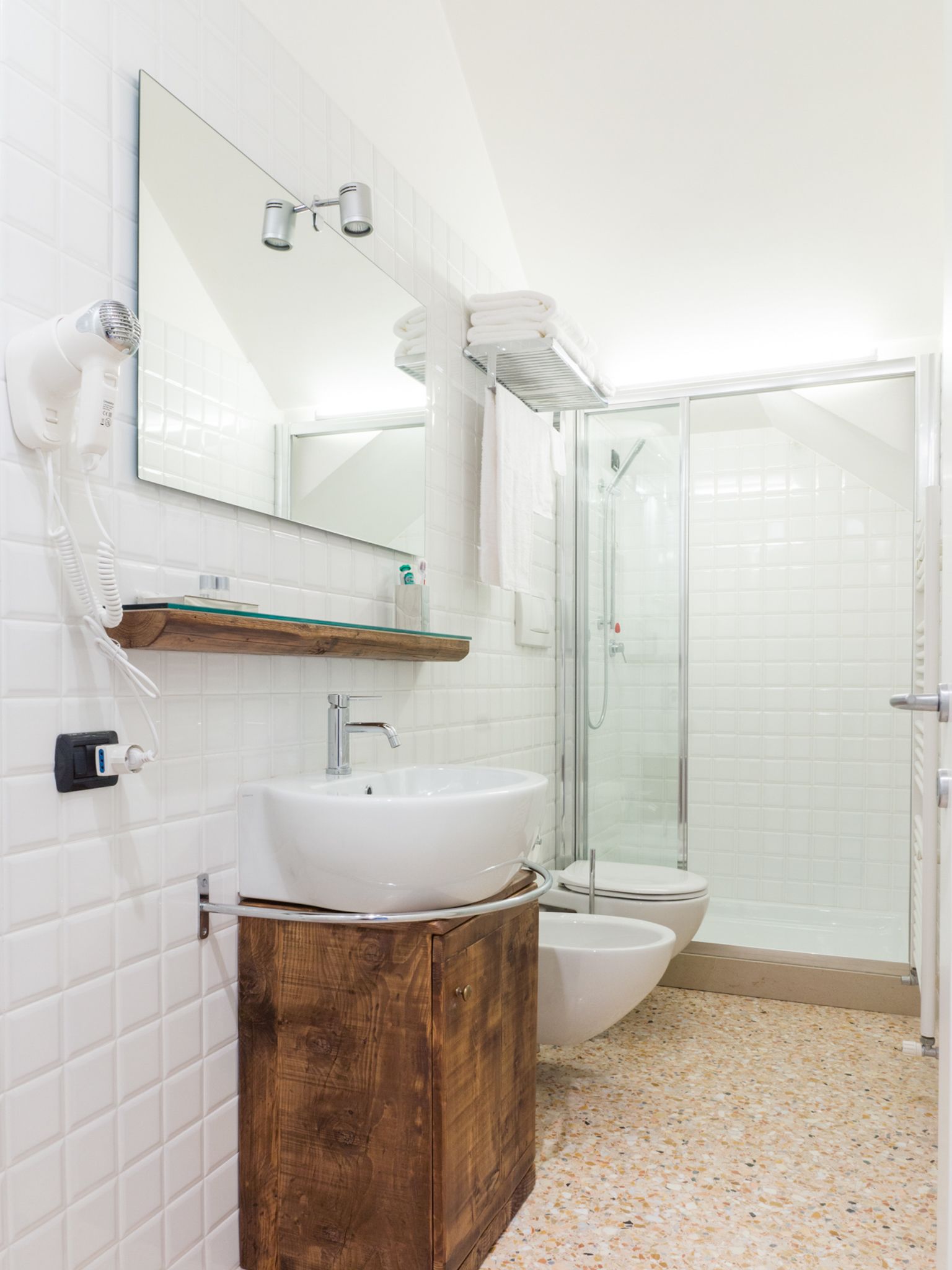 Un baño moderno con lavamanos blanco, tocador de madera, cabina de ducha de vidrio y espejo sobre el lavamanos.