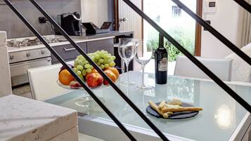 A modern kitchen with a glass table set for a meal, featuring fruits, wine glasses, and a bottle.