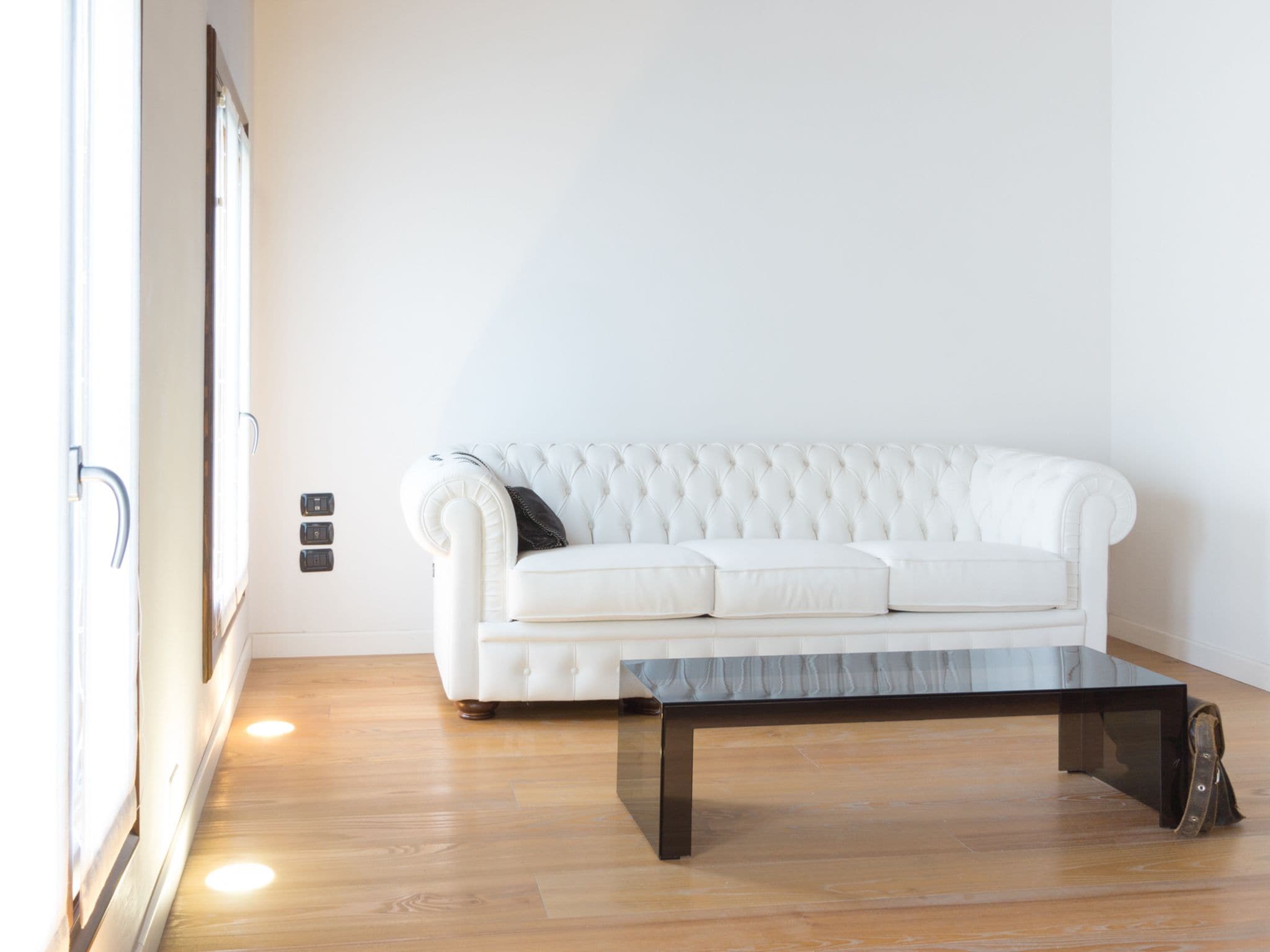 Un sofá blanco con respaldo capitonado y una mesa de centro negra en una habitación con suelo de madera y pared blanca.