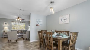 Un coin repas avec une table en bois dressée pour quatre personnes, entourée de chaises en bois. On y trouve un ventilateur de plafond, une fenêtre avec des rideaux et un mur orné d’un tableau encadré.