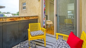 Un balcón con una silla amarilla y un cojín rojo, una mesita y un ventanal de vidrio que da a un espacio interior.