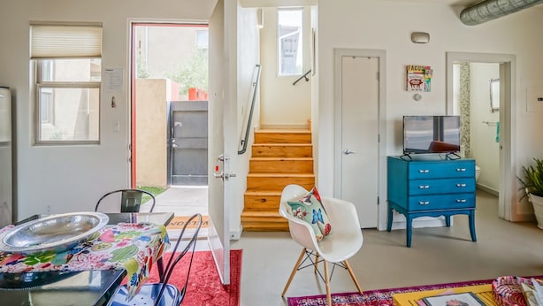 Una habitación con una escalera de madera, un aparador azul, una silla blanca y una mesa con mantel colorido.