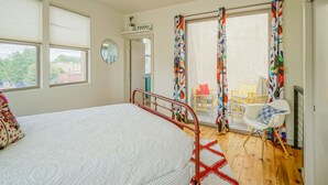 Un dormitorio con cama, ventilador de techo, ventana con persianas, puerta y vista a un patio exterior con sillas y una mesa.