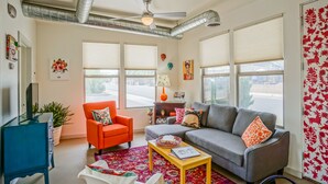 Sala de estar con un sofá gris, un sillón naranja y un aparador azul. Se observan ventanas amplias con persianas, un ventilador de techo y una pared con un estampado rojo.