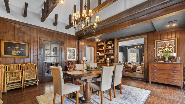 A dining area with a wooden table and chairs, a chandelier, and a bookshelf filled with books and decorative items.