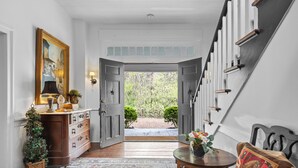 A hallway with a wooden floor, a patterned rug, a wooden bench, a table with a vase, and a door leading outside.