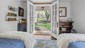 A bedroom with two beds, a white fluffy blanket, a blue patterned rug, a wooden door leading to a garden, and a shelf with books and decorative items.