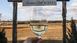 A hand holding a wine glass with the text "Narrow Path Winery" against a backdrop of a sign and a dry landscape.